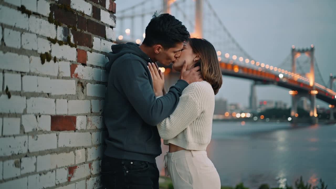 Kissing couple man leaning on brick wall initiating embrace at riverside bridge, hoodie and sweater
