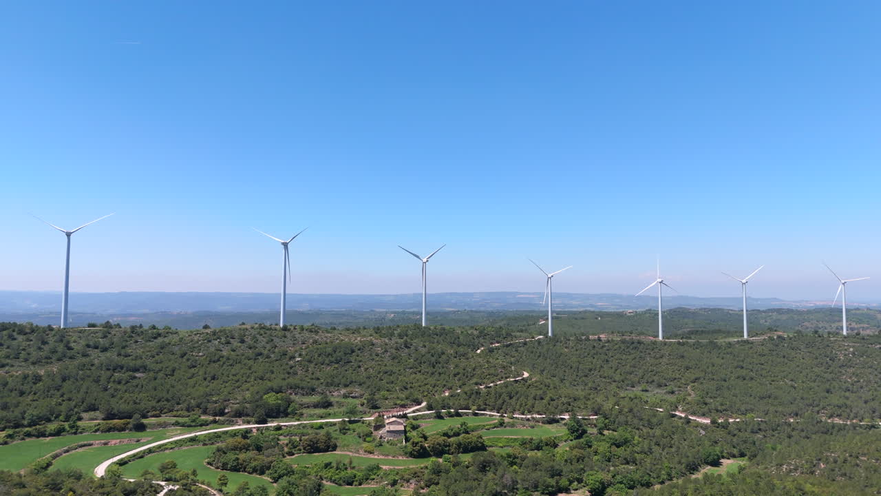 Drone traveling shot flying past wind turbines on forested hills, green farmland, and rural house under a bright blue sky. Renewable energy scene blending technology with nature