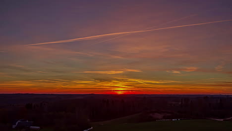 Farbenfroher-Sonnenuntergang-Mit-Dramatisch-Schwebenden-Wolken-Und-Flugzeugspuren-über-Ländlicher-Landschaft-Mit-Häusern,-Wäldern-Und-Feldern