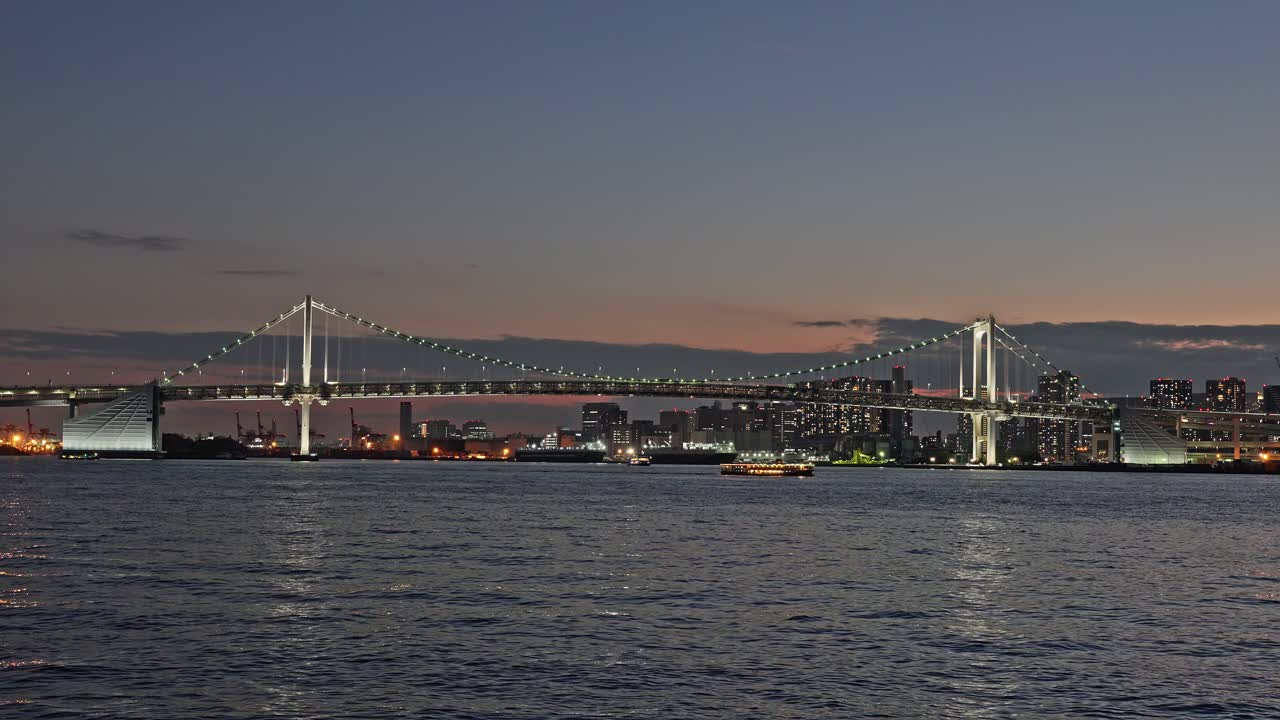 Wide shot of the illuminated Rainbow Bridge and the glowing Tokyo skyline across the dark, rippling bay water