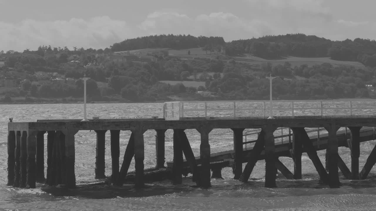 Monochrome wide shot of empty wooden pier, calm water, distant hills, steady camera, natural daylight