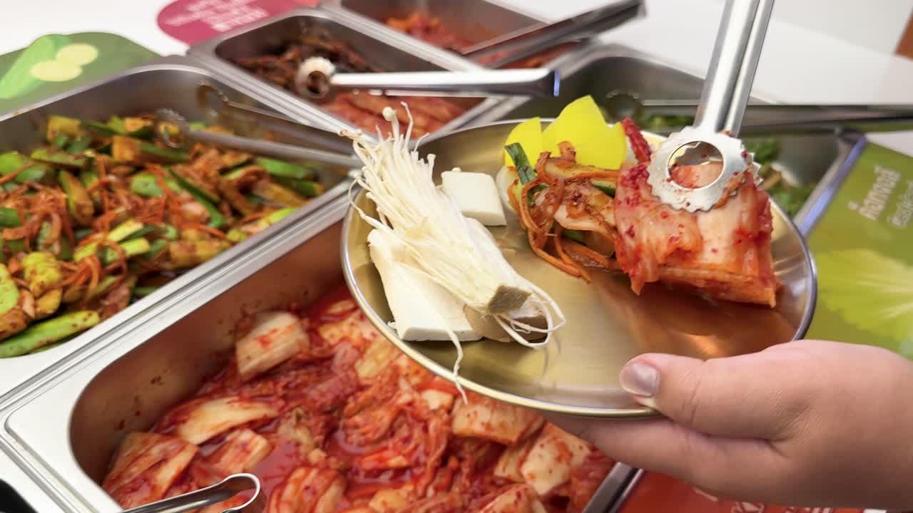 Hand uses tongs to serve kimchi and vegetables onto plate at brightly lit buffet