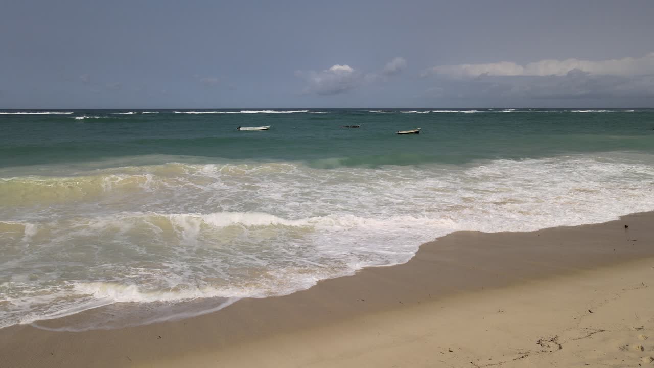 Ondas espumosas do oceano quebrando em uma praia arenosa com pequenos barcos de pesca flutuando em água turquesa