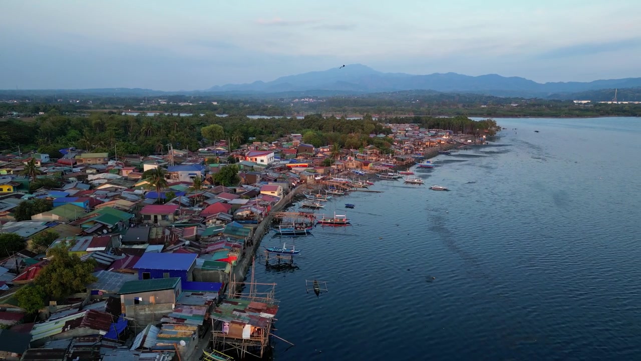 Rekaman drone orbit lambat di sekitar Teluk Batangas di Wawa, Batangas, Filipina, menyoroti rumah-rumah tepi air, perahu-perahu yang ditambatkan, dan punggung bukit pegunungan yang jauh