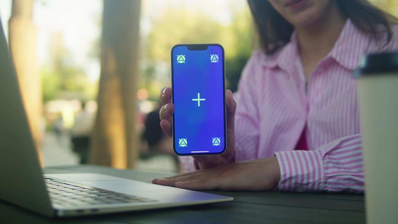 Woman hand holding phone with chroma key screen in front of laptop outdoor. Smartphone with green screen close-up