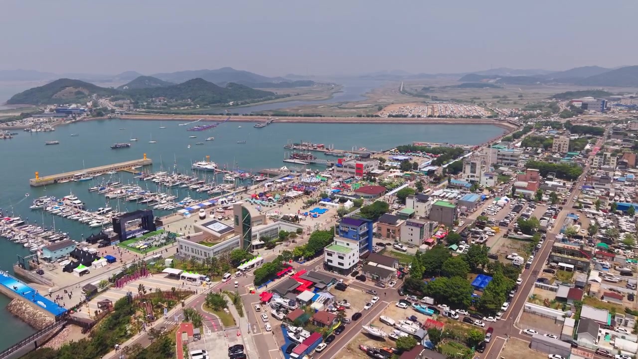 Toàn cảnh bay chậm từ trên cao qua Cảng Jeongok và bến du thuyền trong Lễ hội Thuyền buồm Hwaseong với thị trấn ven biển có đê chắn sóng và bờ sông đông đúc ở Hàn Quốc