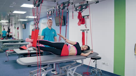 young female is doing exercises with special ropes in rehabilitation center. physiotherapist supports his client during workout to recover after the injury in the modern fitness gym.
