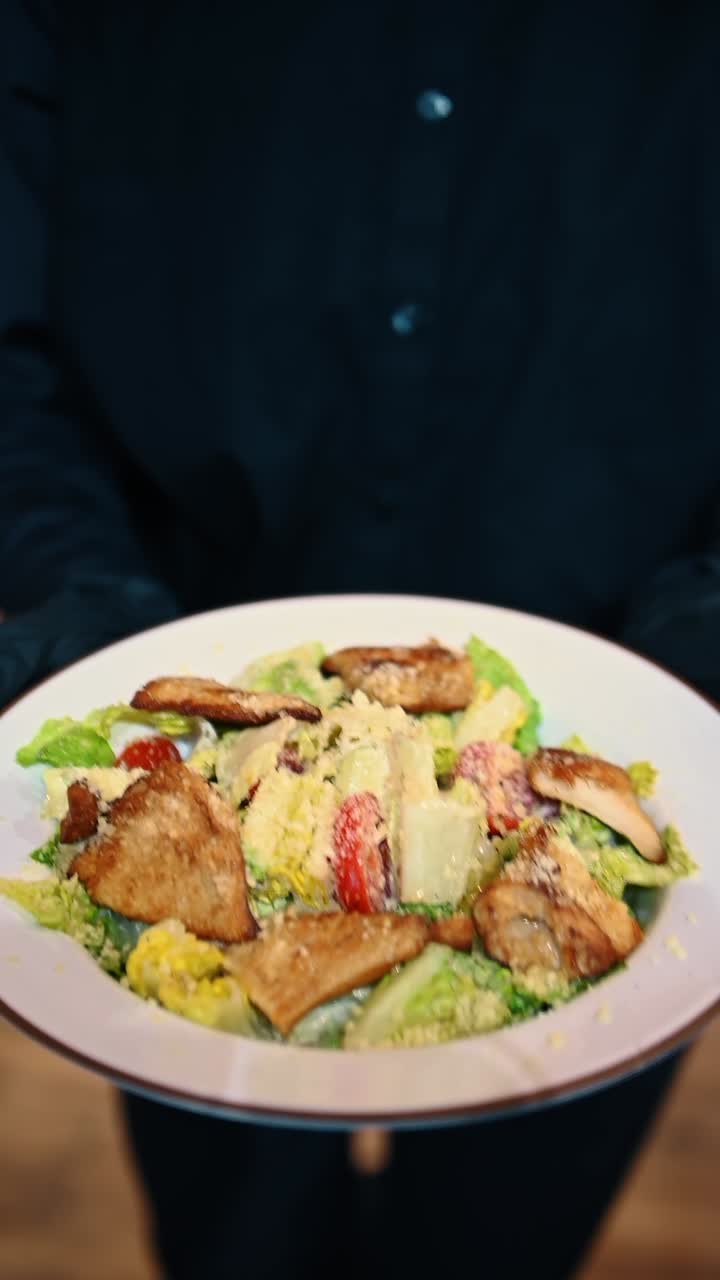 Mani che reggono un piatto di insalata Caesar con pezzi di pollo grigliato. Insalata fresca con lattuga romana, pomodorini, crostini e parmigiano, servita su un piatto bianco.