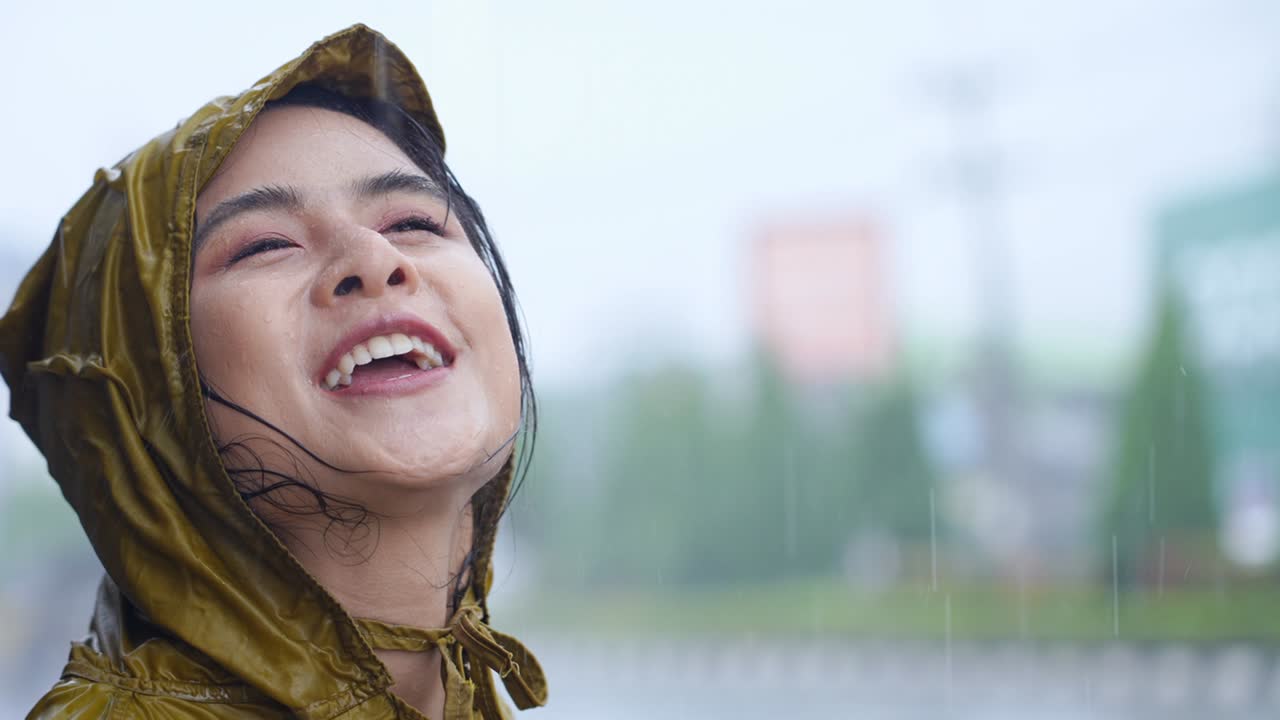 young asian woman wear yellow raincoat enjoy pouring rain standing on the road side, looking up to the sky while rain drops to her face smiling, rainy season weather climate, positive expression