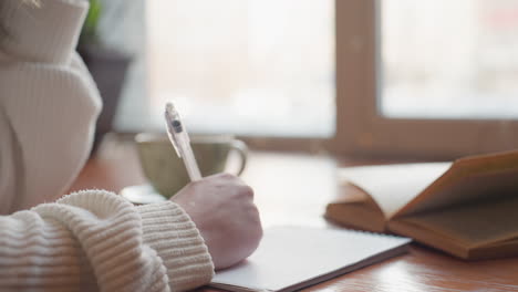 close up of fair skin person writing from novel into notebook with light streaming through window onto wooden table creating peaceful indoor setting ideal for study journaling or creative reflection