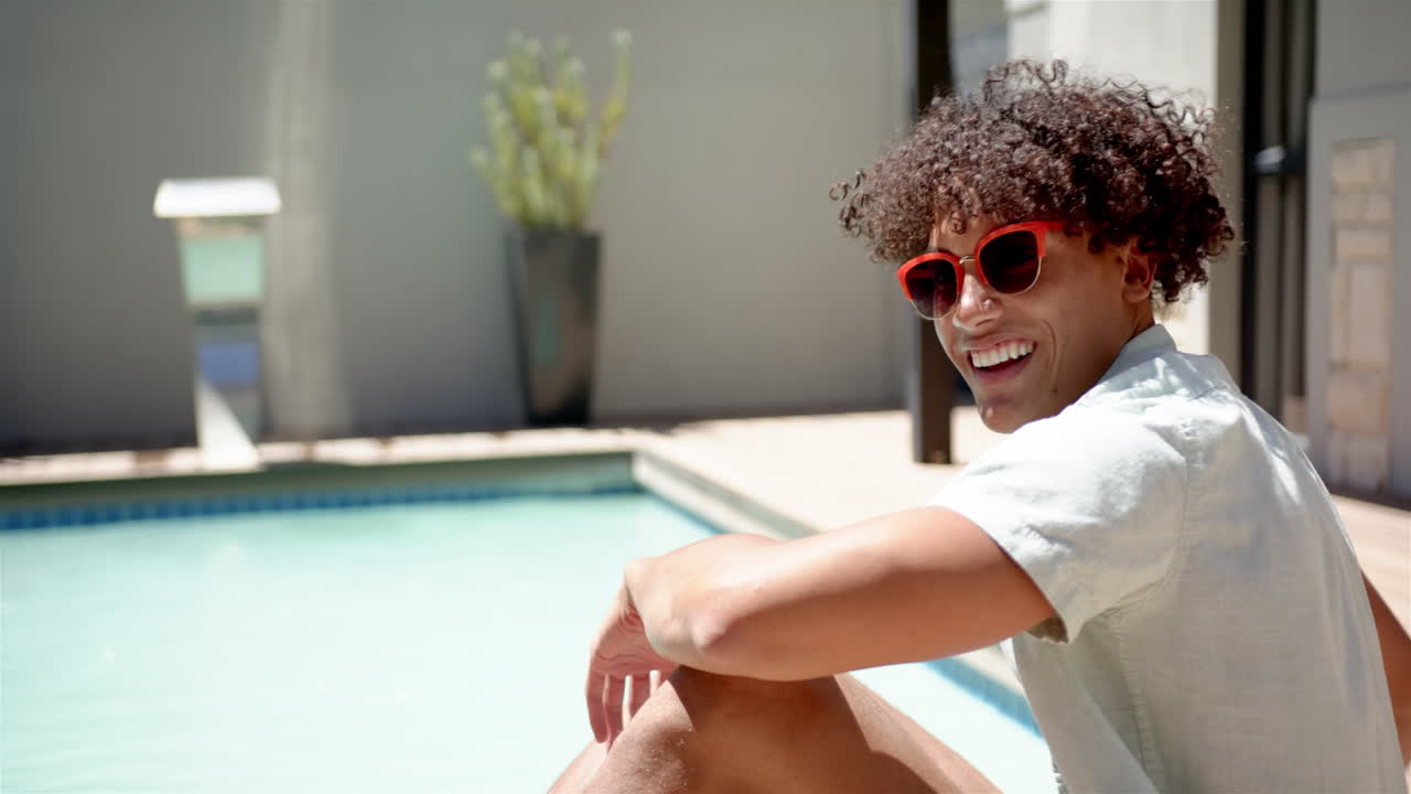 Smiling man wearing red sunglasses sitting by pool, enjoying sunny day, copy space