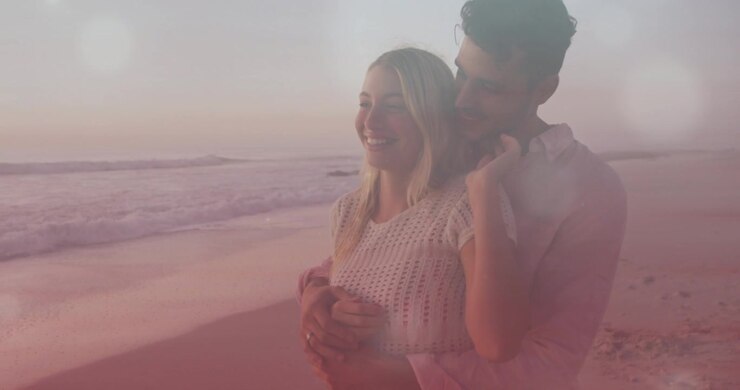 Uomo che si avvicina e avvolge la donna da dietro, chinandosi sorridendo verso l'orizzonte, onde e bagliore dell'obiettivo creano un'atmosfera romantica.