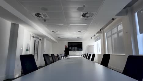 businessman walking in a modern office around a long meeting table with many black chairs and a tv screen