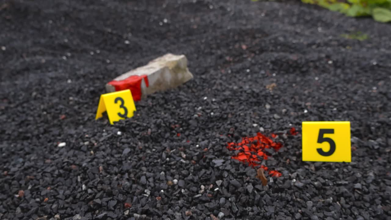 Yellow colored police line Crime scene tape in front of numbered crime markers that are placed next to evidence like a bloody brick or rock and blood spatter or stain on gravel outdoors ground at day