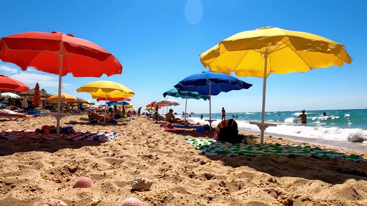 Una Vivace Giornata Estiva in Spiaggia con Ombrelloni Colorati e Conchiglie Lungo la Riva, Perfetta per il Relax e per Godersi le Onde Sotto un Cielo Azzurro Brillante