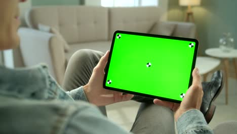 vista por encima del hombro joven en casa sentado en una silla usando con pantalla de maqueta verde tableta computadora en modo horizontal. hombre usando dispositivo de pantalla táctil, navegando por internet