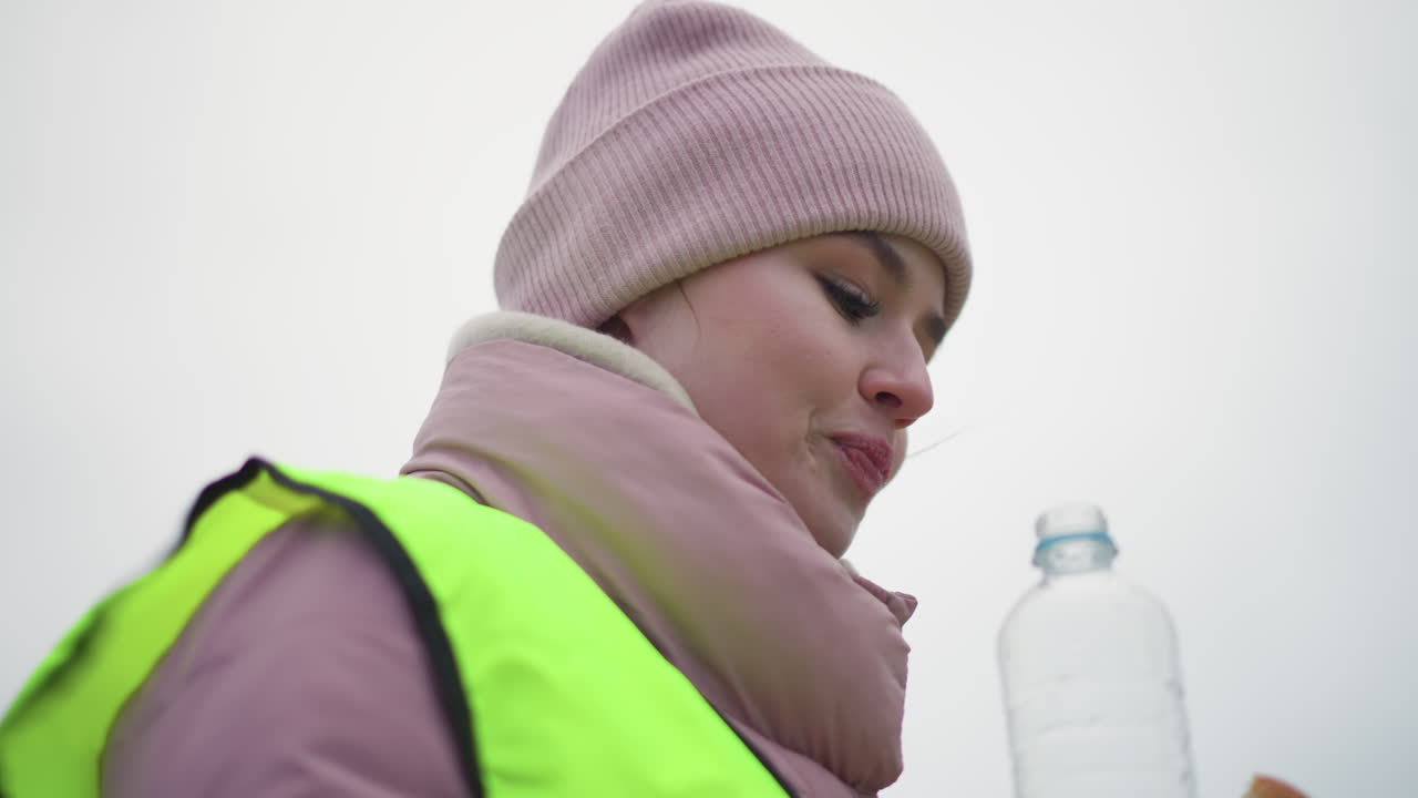 Young female worker in neon reflective safety vest and pink beanie smiling and looking up during outdoor winter work, showing confidence, positivity, and engagement in conversation in cold weather