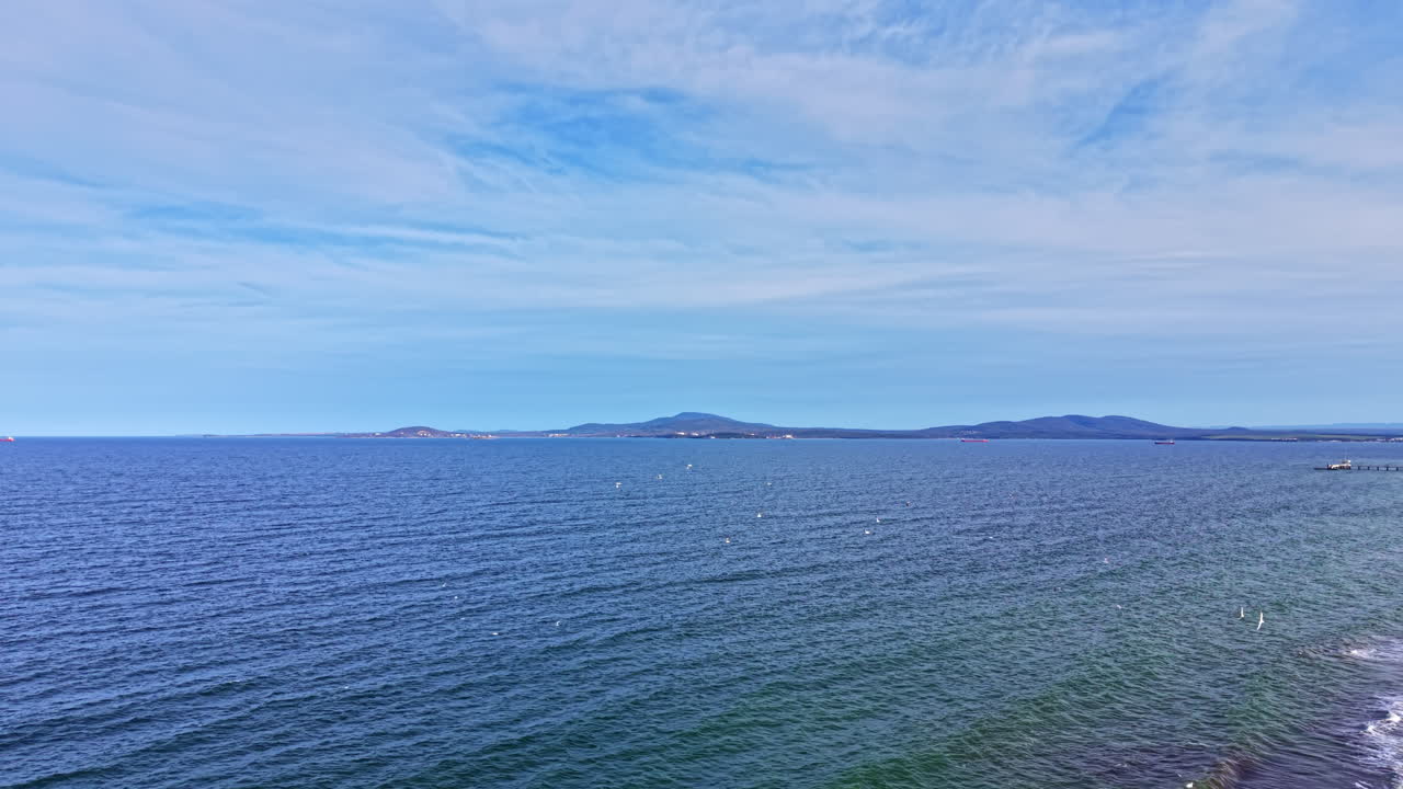 Le onde lambiscono la riva mentre gli uccelli sorvolano un mare calmo vicino alla costa nella luce del pomeriggio, con le colline in lontananza lungo l'orizzonte.