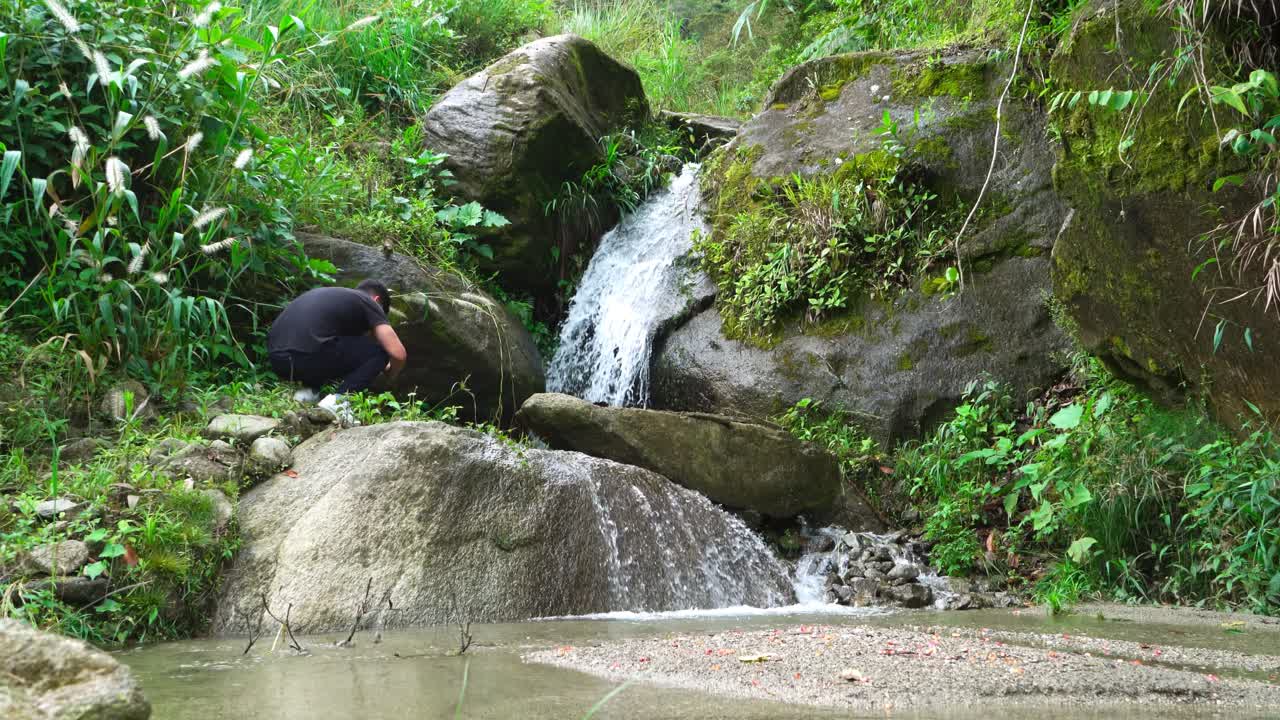 A man photographs a small stream and waterfall in a green forest setting. Ideal for editorial use depicting outdoor exploration and nature photography.
