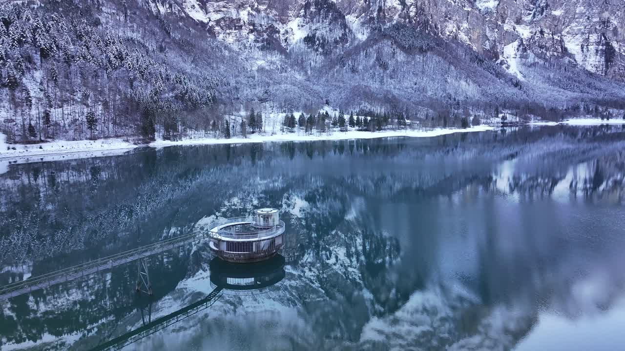 Aerial view of the Klöntalersee lake in Glarus, Switzerland, showcasing a circular water intake structure surrounded by tranquil, reflective waters and snowy alpine scenery.