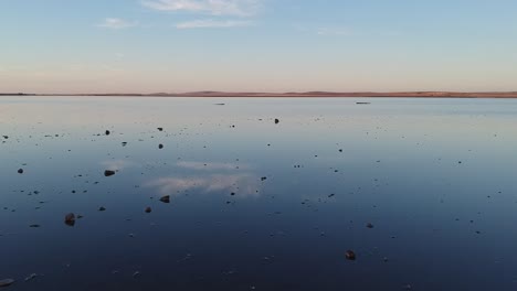 Volando-Bajo-Sobre-El-Lago-Rosa-Y-Lochiel-En-El-Sur-De-Australia,-Con-Rocas-En-El-Borde-Del-Agua-Que-Conducen-A-Nubes-Que-Se-Reflejan-En-El-Agua-Quieta