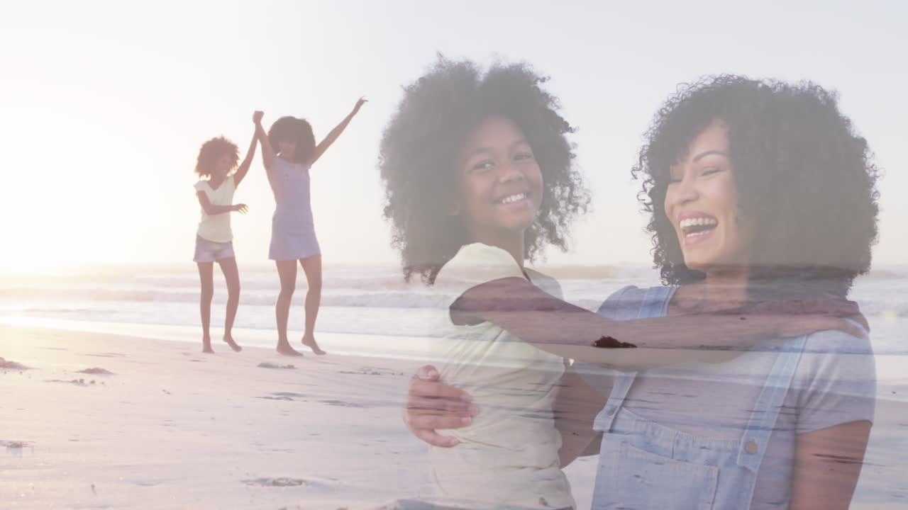 compuesto de una feliz madre y hija afroamericanas abrazándose y levantando los brazos caminando por la playa.