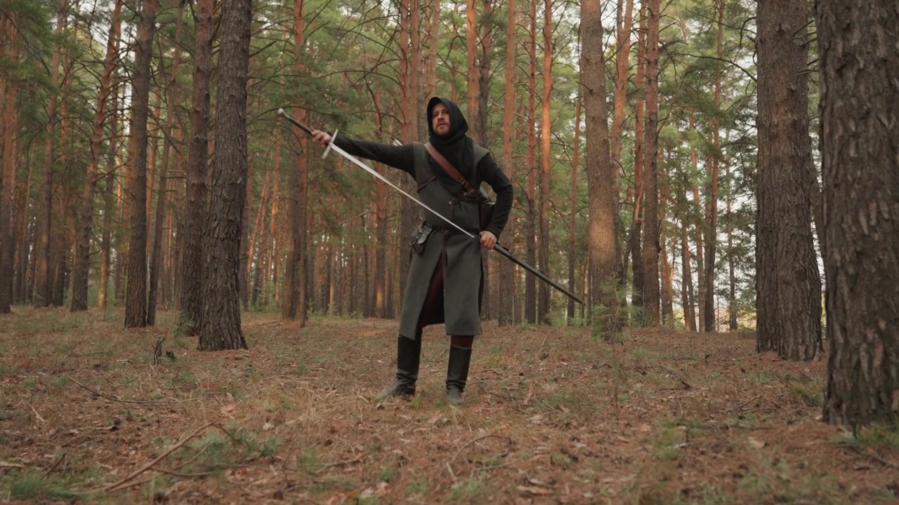 Uma figura solitária e encapuzada avança por um bosque de pinheiros maduros, apoiando um cajado a cada passo firme sobre um terreno irregular coberto por agulhas de pinheiro, galhos e cascas, sob uma luz do dia suave e difusa.