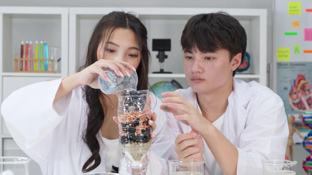 Dos estudiantes con batas de laboratorio vierten agua en un filtro casero durante una clase de ciencias
