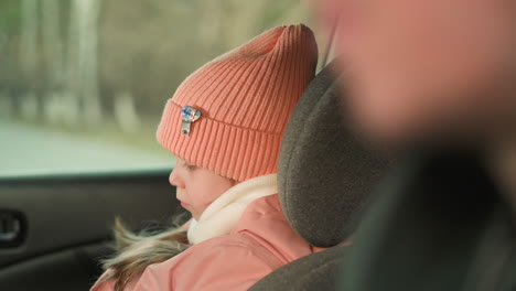 a close-up of a young girl dressed warmly in a pink beanie and jacket, sitting quietly in a car. her serene and calm expression, highlighting the softness of her winter attire and the peaceful moment