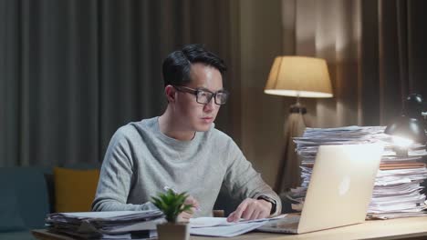 아시아인 남자가 노트북을 보고 집에서 문서와 함께 작업하는 동안 메모를 취하고 있습니다.