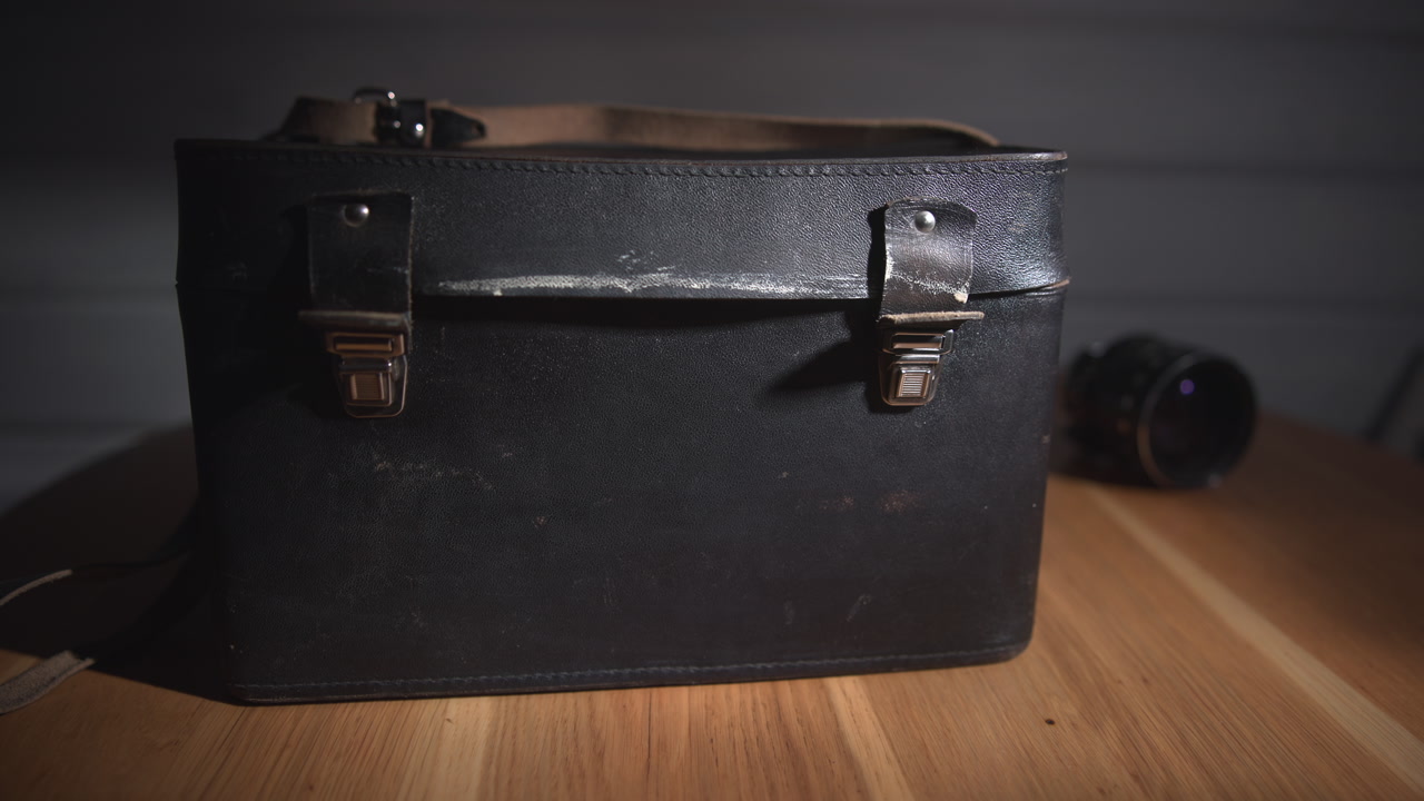 Worn Leather Camera Case On Wooden Table, Dim Studio Lighting, Scuffed Metal Clasps, Vintage Lens Beside Case, . Media