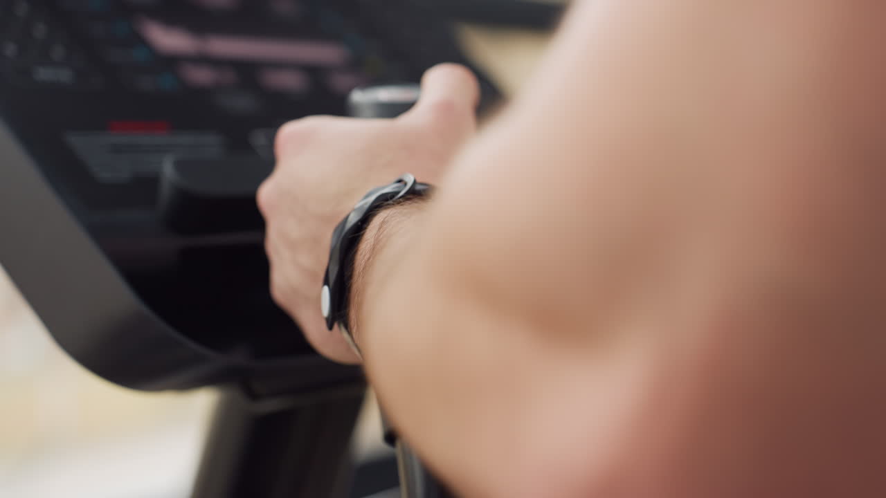 Hand view of gym trainer gripping treadmill handle while adjusting console on elliptical trainer in modern fitness center with large windows forearm strength display intensity and control focus