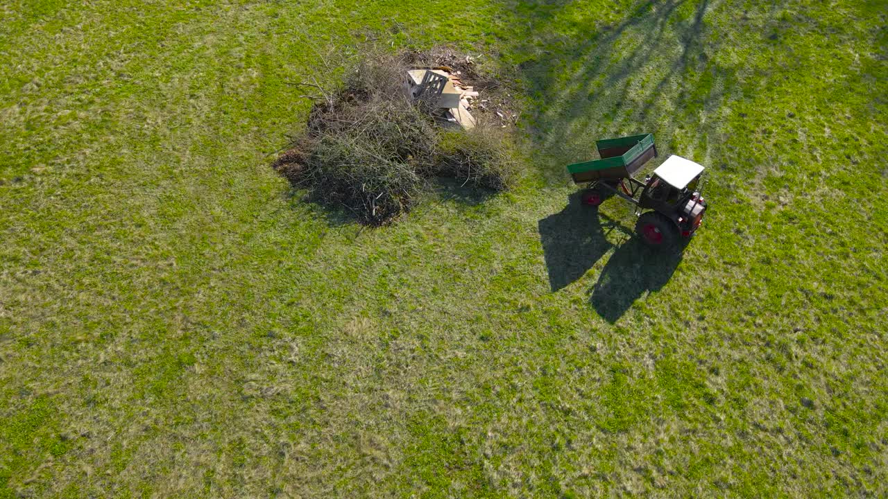 Aerial drone footage of a small vintage and retro beautiful looking tractor working in a green grassy farm field in the countryside. The tractor vehicle is carrying branches and leaves, sunny day time