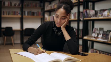 atractiva joven concentrada sentada junto al escritorio y tomando algunas notas en su cuaderno para la universidad en la biblioteca moderna, preparándose para los exámenes