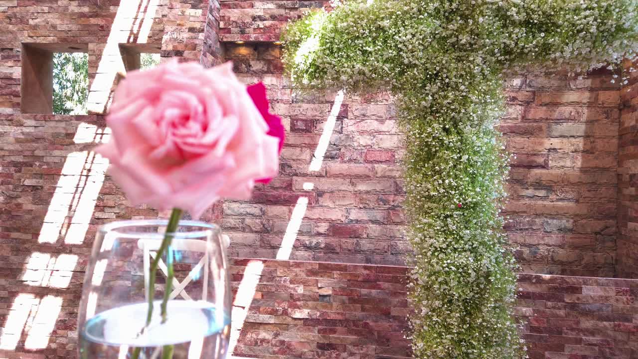 A close-up of a pink rose in a glass with a wall covered in flowers and a cross-shaped structure in the background.