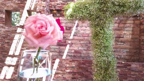 a close-up of a pink rose in a glass with a wall covered in flowers and a cross-shaped structure in the background.