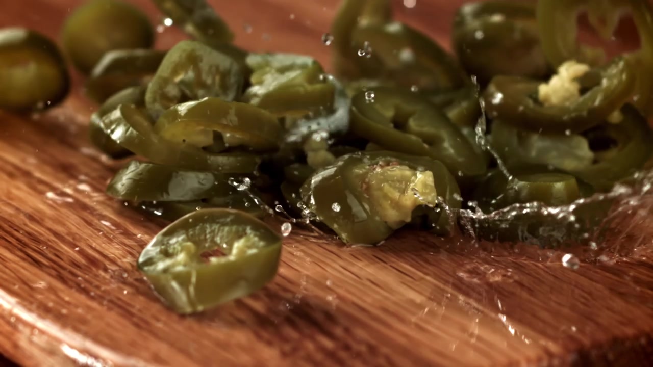 Slow motion av skivade inlagda jalapeños som faller ner på en våt träskiva.