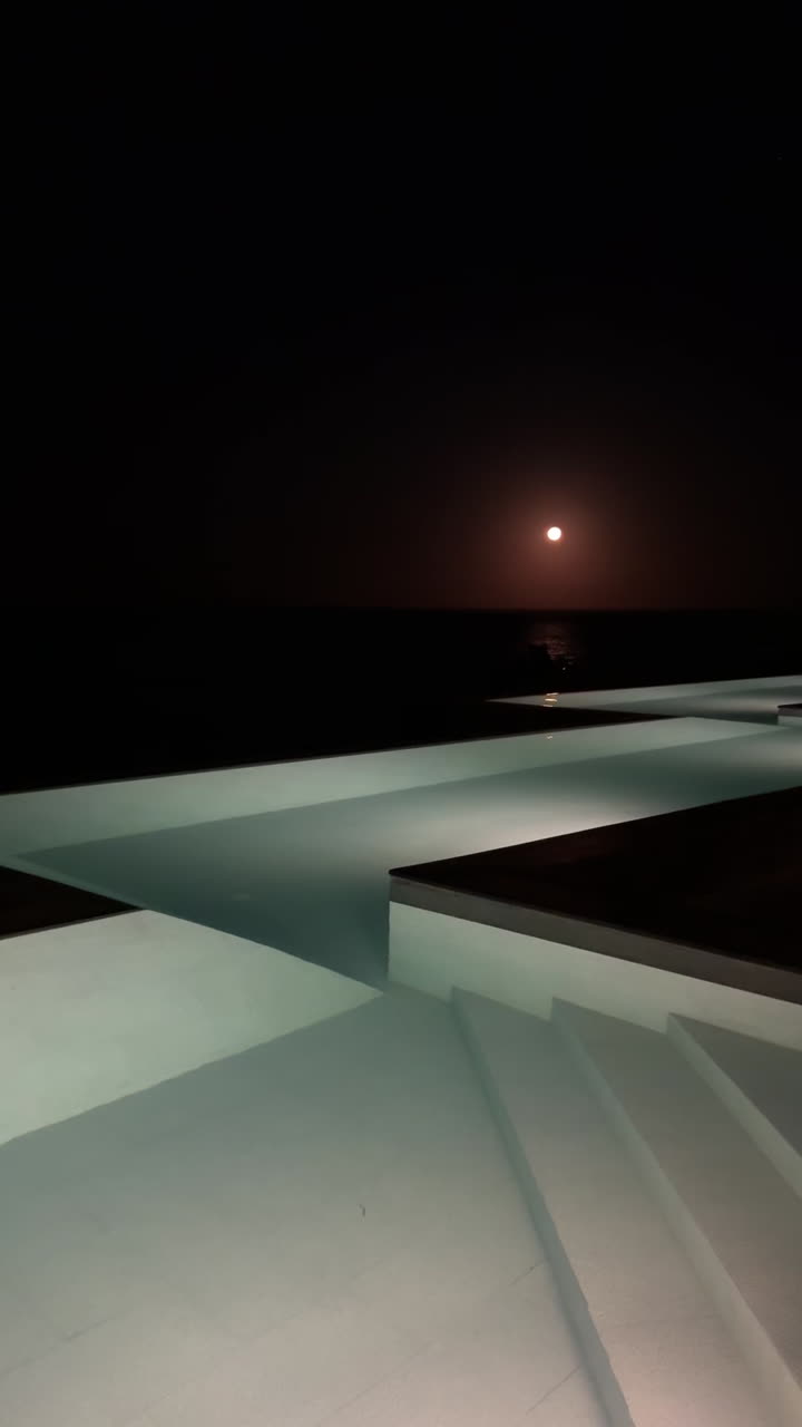 Nighttime view of a swimming pool overlooking the ocean with a full moon in the sky