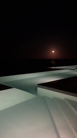 nighttime view of a swimming pool overlooking the ocean with a full moon in the sky