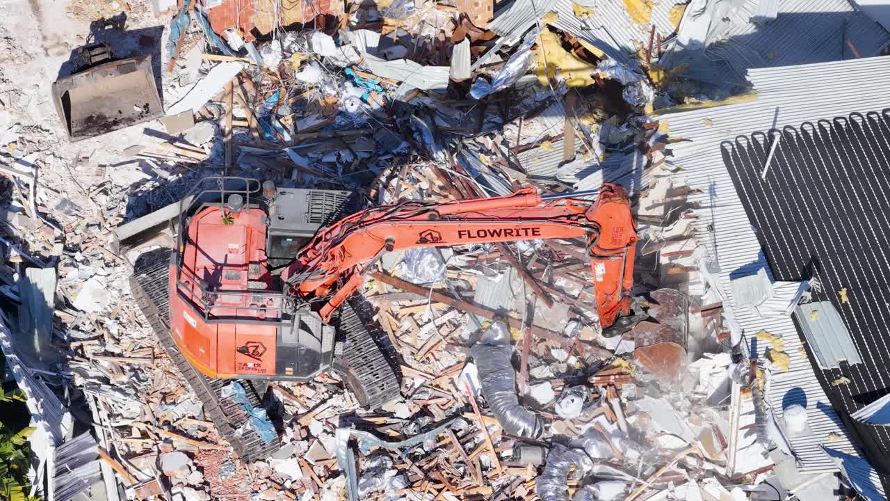 Aerial view of an orange excavator actively demolishing a house, surrounded by debris and construction materials, under bright daylight with clear shadows