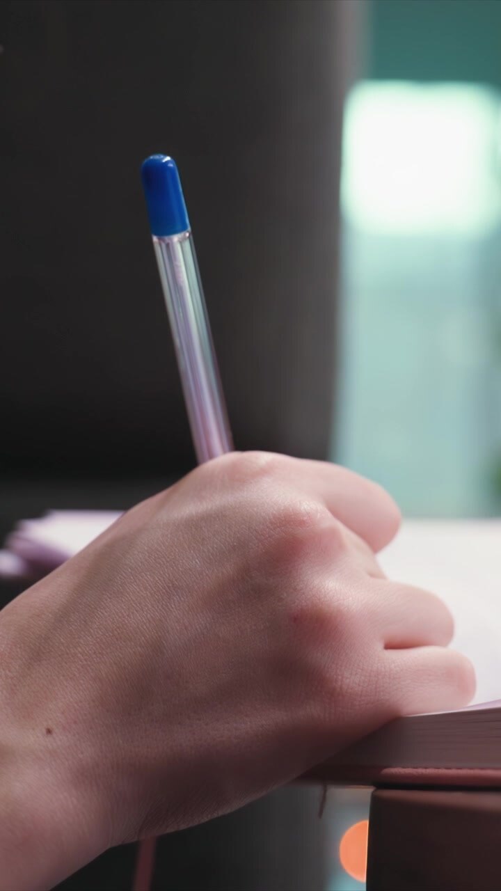 Il primo piano mostra la mano di una persona che impugna una penna trasparente con tappo blu sopra un libro aperto o un foglio. La scena si concentra esclusivamente sulla texture della pelle e sul movimento implicito nel tenere e posizionare la penna per prendere appunti.