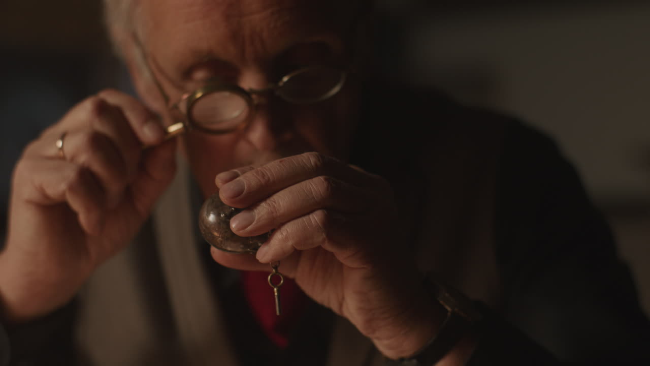 Older passionate man evaluates an old silver skull watch in his office at night