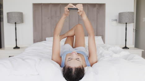 Happy-biracial-woman-lying-on-bed-using-smartphone,-slow-motion