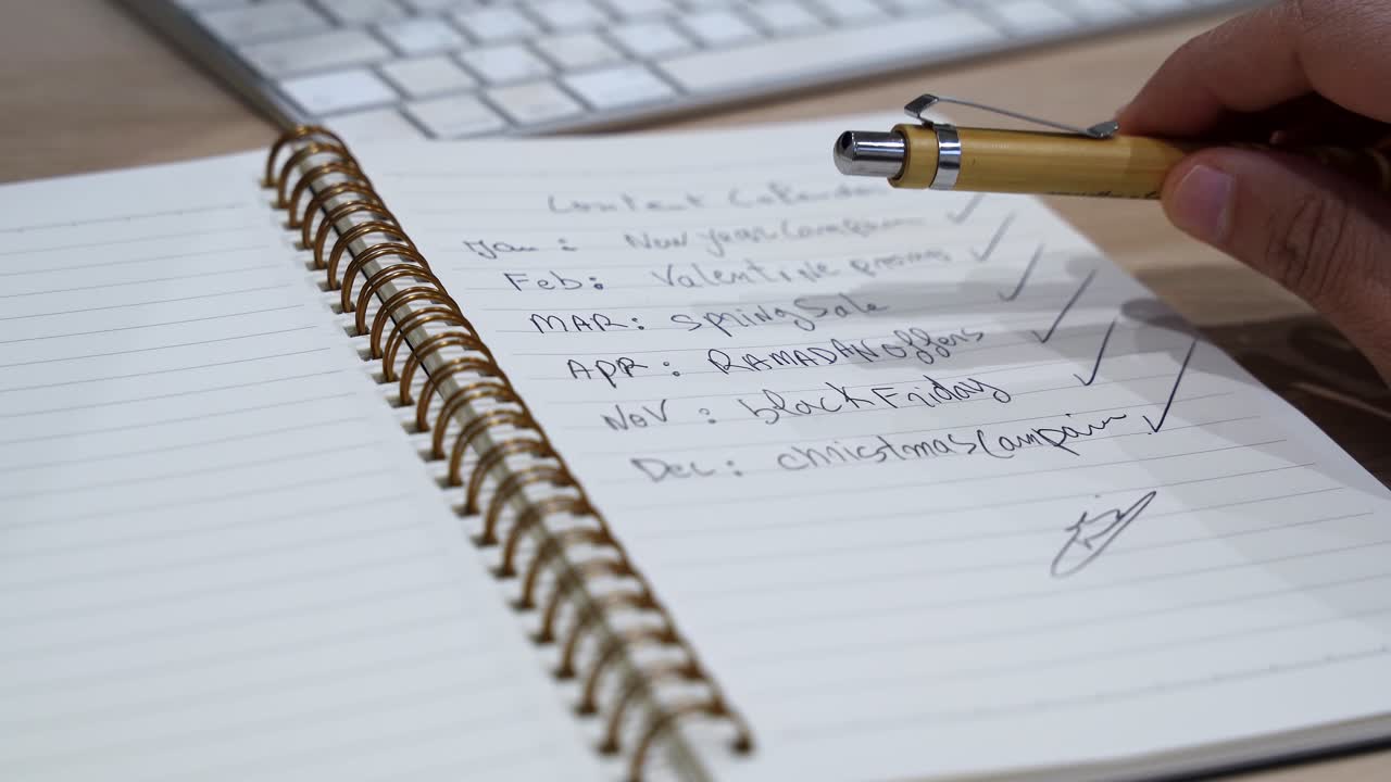 Close-up of a hand holding a pen above a handwritten content calendar in a spiral notebook on an office desk, with keyboard in soft indoor light and shallow depth of field