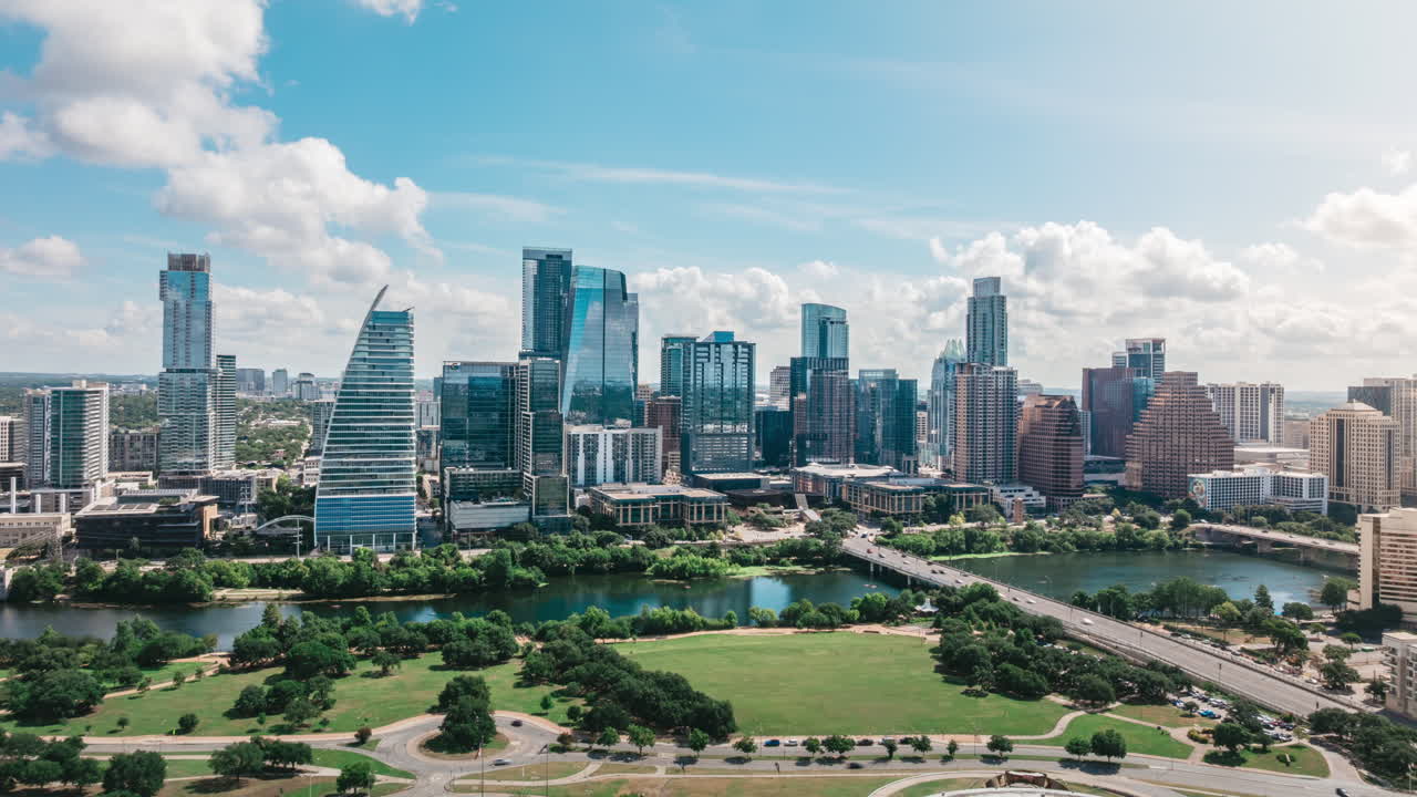 Drone hyperlapse orbits Auditorium Shores, smoothly panning across Austin Texas skyline and Ladybird Lake. Busy summer morning scene with vibrant urban life, water reflections blue skies with clouds