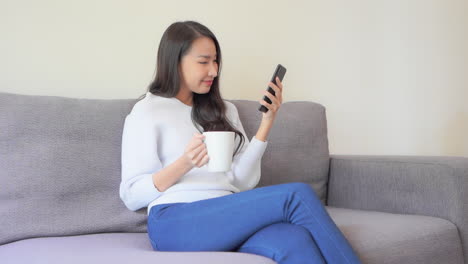 asian woman with cup of tea sitting on a couch and watching phone with happy face expression