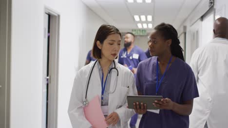 video of two diverse female doctors walking in busy hospital corridor, looking at tablet and talking