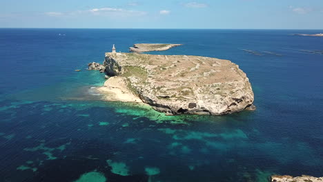 a flying upward drone shot of a small, uninhabited island, surrounded by the blue ocean with turquoise patches in malta on a sunny day.