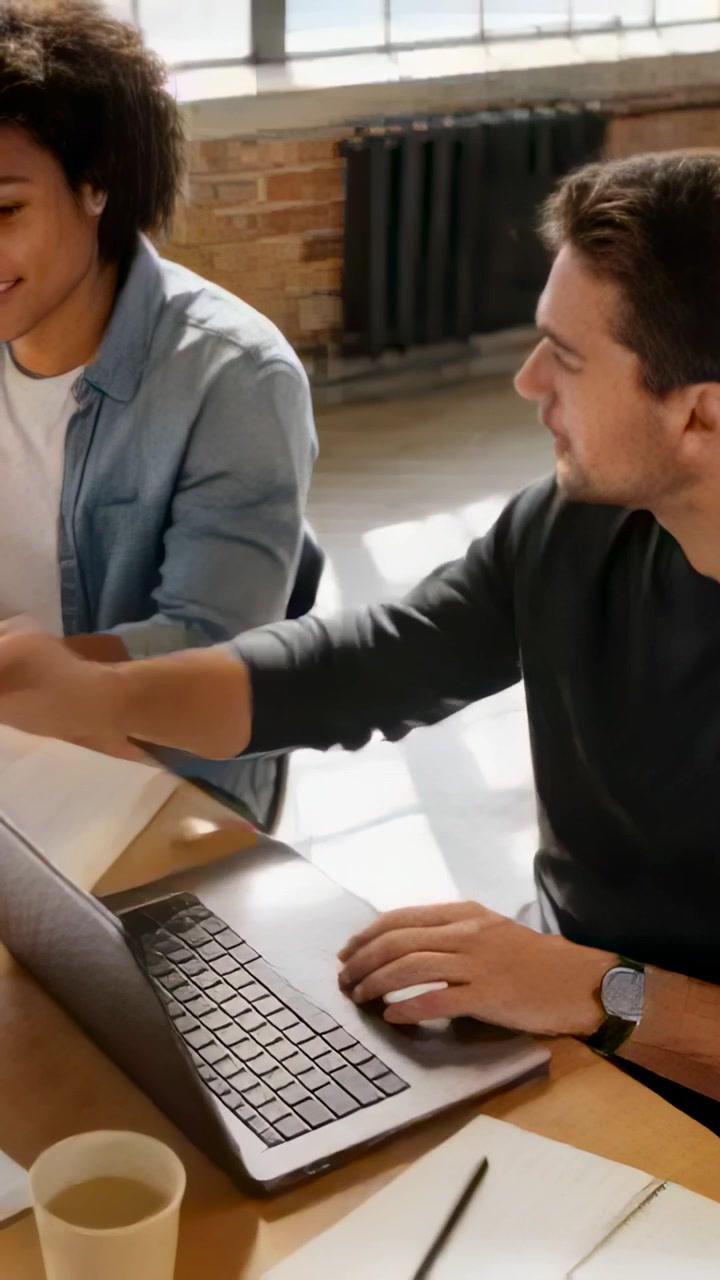 Video verticale: Uomo in maglietta scura che indica e usa il laptop, mentre una donna in camicia di jeans controlla documenti in un loft.