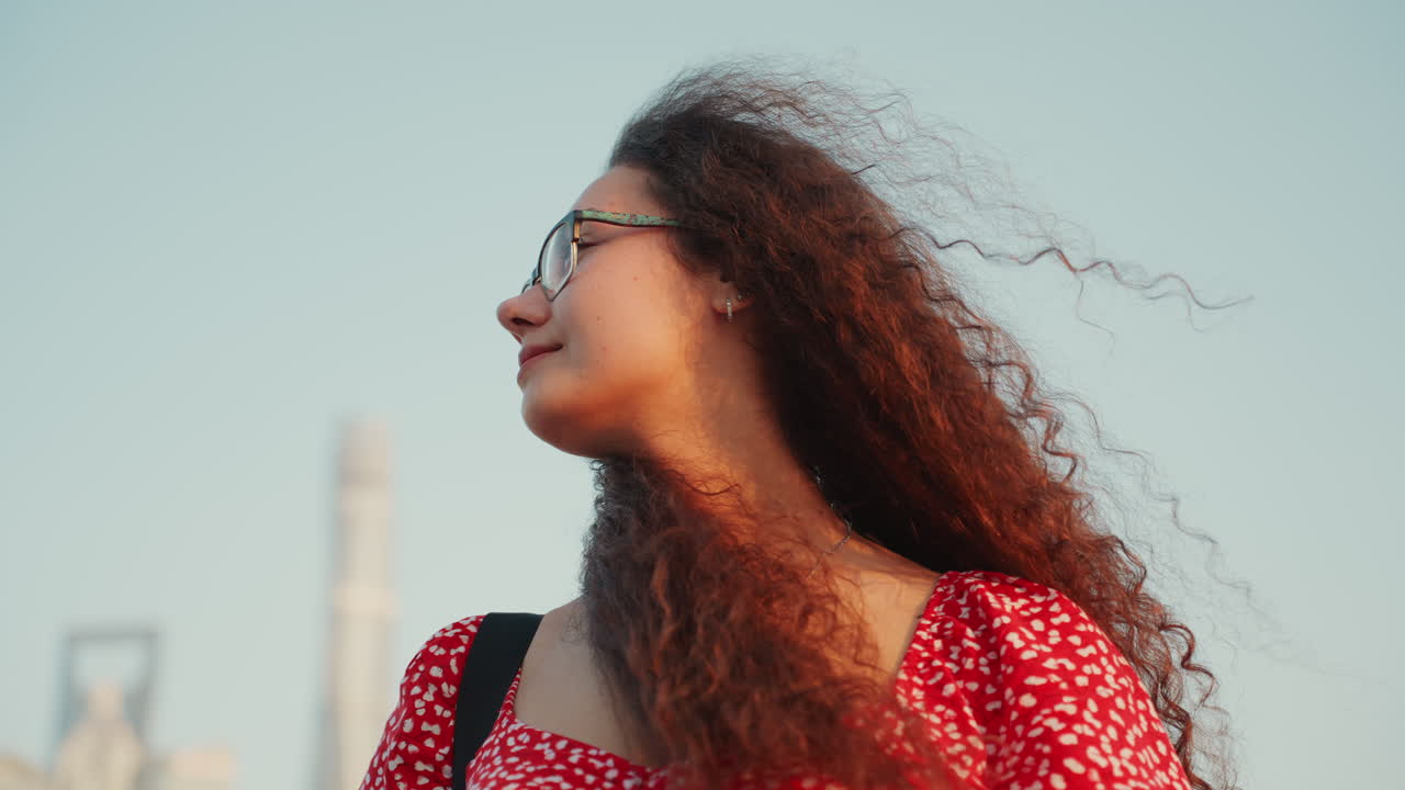 Closeup multiracial woman with glasses, curly hair against sunset skyline contemplative expression, windswept curls, red dress, soft golden light, rooftop towers in background, candid portrait